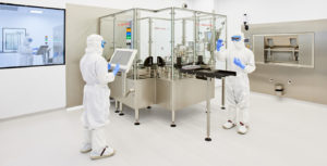 Two people completely covered in personal protective equipment operate an automated vial-filling machine in a 503B pharmacy