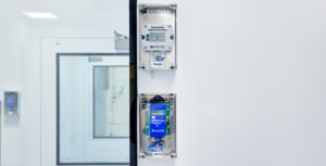 A wall-mounted sensor monitors the temperature and relative humidity while another wall-mounted sensor below it monitors the differential pressure of a cleanroom within a 503B pharmacy