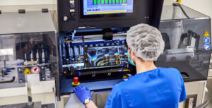 A person in blue scrubs, hair net, blue surgical face mask, purple nitrile gloves and blue shoe covers inspects filled amber vials using a semi-automated vial-inspection machine